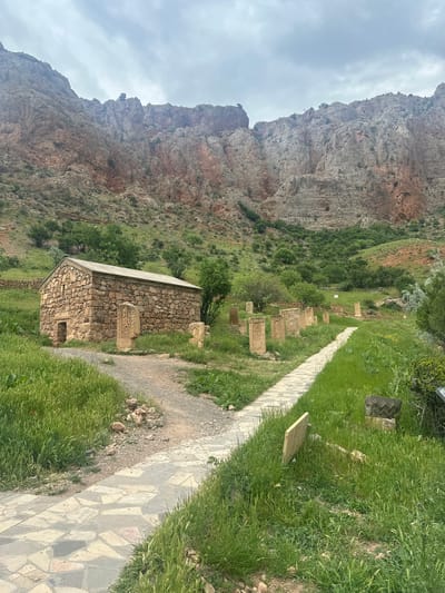 This image depicts a historical site located in Amaghu, Armenia, under an overcast sky. The scene is dominated by a rugged mountainous backdrop composed of grey and reddish-brown rock formations, with sparse green vegetation on its lower slopes.
In the mid-ground, a small, rectangular stone building stands on the left. It is constructed from irregular, light brown and grey stones and features a dark grey, slightly pitched roof. A recessed, arched doorway and a small window are visible on its facade.
To the right of the building and extending into the background, a series of upright stone monuments, likely Khachkars (Armenian cross-stones), are arranged within a vibrant green grassy field. These monuments are made of light-colored stone and display carved patterns.
Two pathways traverse the scene. A dirt path curves towards the stone building from the foreground. Parallel to this, a paved pathway, constructed from irregularly shaped grey and beige flagstones, extends straight back into the distance, bordered by the grassy area and several more monuments on its right side.
The ground is covered with lush, tall green grass, interspersed with small trees and shrubs, particularly evident in the mid-ground. No people are visible in the scene. The lighting is soft and diffused, indicative of a cloudy day.