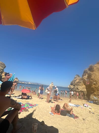 The image depicts a crowded beach scene under a clear, bright blue sky in Lagos, Portugal, specifically identifiable as one of the beaches within the city's bay, such as Praia da Batata, due to the characteristic golden sandstone cliffs, sea stacks, and the distant view of the city coastline.

In the foreground, the upper left quadrant is dominated by the underside of a large beach umbrella, segmented into red and yellow panels, providing shade. Below this, a person is seated on the sand, facing the water, with their left shoulder and tattooed arm visible. A white disposable cup with a red straw is positioned in the sand near their feet, within the umbrella's shadow.

The central and mid-ground sections of the image reveal a sandy beach populated by approximately 30-40 individuals. Most are in swimwear. Actions observed include sunbathing on various patterned towels (floral, striped), standing, walking, and wading or swimming in the calm, blue-green ocean. Notable individuals include a person lying face down on a striped towel with visible Adidas slides in the lower right, another person lying on their side in the mid-ground, and a man in a white straw hat standing near the water's edge. To the left, a man wearing a cowboy hat is seen standing near a blue beach umbrella, holding a beverage container. A small red beach umbrella provides shade for a person resting beneath it in the mid-ground.

The environment is characterized by golden-hued, eroded sandstone cliffs and distinct rock formations or sea stacks bordering the beach on both the left and right sides. Sparse green shrubbery grows on the cliff faces. A warning sign, predominantly red and yellow, is affixed to the cliff face on the right. In the background, the open bay extends towards a distant coastline featuring a city or town with white buildings under the clear sky.