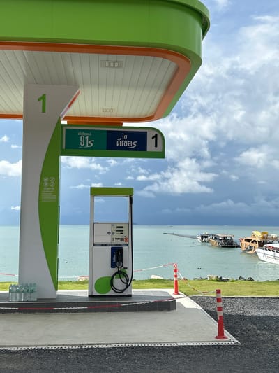 The image depicts a modern, brightly colored gas station canopy with green and white accents, situated in Ratsada, Thailand. The canopy covers a single gas pump, which is white with green circular accents and a black hose. To the left of the pump, a large white pillar displays the number "1" in green. Below this pillar, several bottles of water are neatly arranged. A green sign above the pump indicates fuel types in Thai script.
The gas station is positioned adjacent to a body of water, likely the Andaman Sea, given its location in Thailand. The water is a tranquil blue-green, and several boats, including speedboats and a larger vessel with a wooden hull, are docked or moored in the distance. A wooden walkway extends into the water. The shoreline is lined with green grass and a low wall. The sky is partially cloudy, with dramatic, dark clouds in the distance contrasting with lighter, scattered clouds overhead. Red and white stanchions with chain barriers mark the perimeter of the paved area. The overall impression is one of a clean, well-maintained, and picturesque service station by the sea.