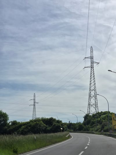The image shows a view of power lines and pylons against a cloudy sky, alongside a road and treeline. The pylons are metal lattice structures. Streetlights are visible among the trees. The road is asphalt with a white line marking the edge. Based on the visible signage and visual style, the location is likely in Sanem, Luxembourg. There are no people visible in the image.