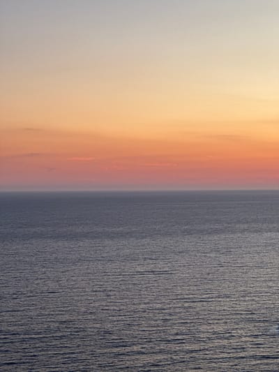 The image depicts a serene seascape at sunset, viewed from Glavatičići, Montenegro. The sky transitions from a pale, warm yellow at the top to a soft orange and pink closer to the horizon. The sea, a deep, muted blue-grey, exhibits a gentle ripple pattern, reflecting the subdued light of the setting sun. There are no people, objects, or structures visible in the frame, emphasizing the vastness and tranquility of the natural environment. The horizon line is sharp and clear, separating the sky from the ocean.