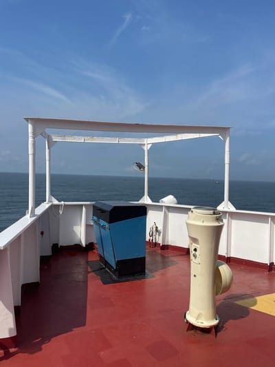 The scene depicts the forward deck of a marine vessel situated off the coast of Orimedu, Nigeria, under a clear blue sky with sparse, wispy clouds. The deck surface is coated in a weathered red-orange anti-slip paint, with a segment of yellow marking visible in the lower right corner. White-painted metal bulwarks and railings enclose the deck area.
A white, open-frame metal canopy structure, supported by four pillars, is centrally located on the deck, exhibiting minor surface corrosion. A dark, possibly surveillance, device or floodlight is affixed to one of the canopy's forward pillars, oriented towards the sea.
Two primary pieces of equipment stand on the red-orange deck:
1. A freestanding, rectangular equipment housing with a sloped front, painted black on its top and bottom sections, and blue on its central, slanted portion. It features an access panel or lid on its upper surface.
2. A cream-colored, fluted pedestal-style unit, approximately one meter in height, with a flared base and top, featuring multiple control panels or ports on its side. This unit is positioned to the right of the blue and black housing.
The vessel overlooks a calm, dark blue sea, extending to a flat horizon line. Several small, distant maritime objects, identifiable as vessels or buoys, are visible on the water's surface. The deck is devoid of human presence.