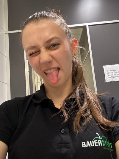 A young woman with brown hair pulled back in a ponytail is taking a selfie in what appears to be a public restroom. She is wearing a black polo shirt with the "BAUER MART Regional" logo on the left chest. She is winking and sticking out her tongue, with her left eye closed and her right eye partially open. The background is a gray wall with white paneling and a sign that reads "Die Toilette ist kein Tatort. Spuren dürfen beseitigt werden." This translates to "The toilet is not a crime scene. Traces may be removed." The lighting suggests indoor fluorescent lighting. The overall impression is a playful, informal moment captured in a utilitarian space. The specific location of Elsenfeld, Germany is implied by the text on the sign.
