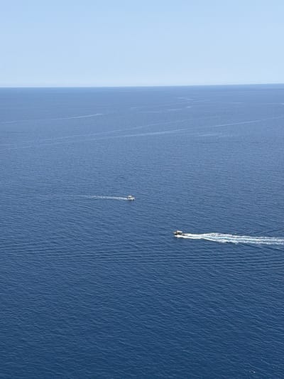This aerial image depicts the open ocean near Lindos, Greece. Two boats are visible, each leaving a wake behind it as it moves across the water. One boat is further away, with a more pronounced wake indicating higher speed. The water surface is textured with small waves. A faint coastline is visible on the left.