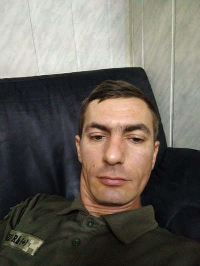 A man is sitting in a dark chair, looking at the camera. He is wearing a dark green shirt with a patch on the left chest. The patch has the letters "WRH" and a symbol. The man has short brown hair and is looking down slightly with his eyes. His skin is light brown and he has some stubble on his face. The background of the image is a light-colored wall with vertical paneling. The image is taken from the chest up. The image is likely taken inside a building.