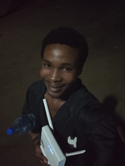 The image shows a young man of African descent, appearing to be in his late teens or early twenties, holding a plastic water bottle with a blue cap in his left hand and a white electronic device, possibly a router or modem, in his right. He is wearing a black shirt. The man is looking directly at the camera with a slight smile. The background is dark and appears to be an outdoor, unlit area. The scene is consistent with the urban environment of Orodo, Nigeria, where such street-level technology and public spaces are common. The lighting suggests it is nighttime or a poorly lit indoor setting.