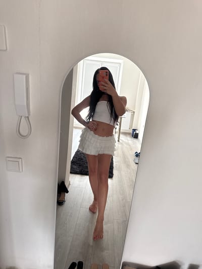 A woman stands in a room, holding a phone and taking a selfie in a large oval mirror. She is wearing a white bandeau top and a white ruffled mini-skirt. Her hair is dark and falls past her shoulders. Her right hand is on her hip and her left hand holds the phone up to take the picture. The floor is light wood with a dark rug visible near her feet. A black high-heeled shoe is on the floor to the left of the rug. To the left of the mirror, on the wall, is a white intercom system with a coiled cord. The room appears to be an apartment in Zapadny, Russia.