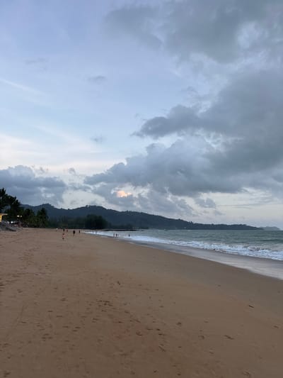 The image depicts a wide, sandy beach in Ban Bang Niang, Thailand, during what appears to be dusk. The sand is a light brown color, with scattered footprints and some small debris. The sea is to the right, with gentle waves rolling onto the shore and white foam at the water's edge. The water itself is a greenish-blue. In the distance, a line of dark green hills and mountains forms the backdrop, covered in trees. The sky is overcast with dramatic, dark gray clouds dominating the upper portion of the frame, interspersed with lighter patches of blue and a hint of warm light near the horizon. On the far left of the beach, there are palm trees and some structures that suggest a resort or a row of shops, with faint lights visible. Several people are scattered across the beach, walking and standing, with a few children playing near the water's edge. The overall scene conveys a sense of tranquility and natural beauty, with a touch of impending weather.