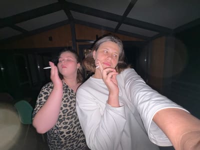 Two young women are standing outdoors at night, possibly under a covered patio or porch. The woman on the left is wearing a leopard-print shirt and is holding a lit cigarette to her mouth with her eyes closed. The woman on the right is wearing a white long-sleeved shirt and is holding a lit cigarette between her fingers, looking to the side. The environment appears to be a residential area in Костоваты, Russia, with wooden structures and dark surroundings.