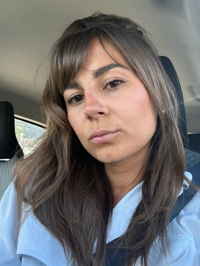A young woman with long brown hair and brown eyes is seated in a vehicle. She is wearing a white shirt and the seat appears to be black. The background outside the window shows a city landscape with buildings and greenery, characteristic of a hilly terrain. The image does not definitively confirm the location as Mougins, France, but the landscape visible through the window suggests a Mediterranean setting.