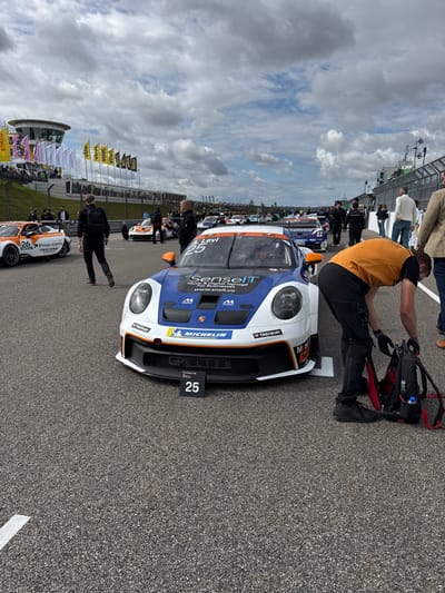 The image depicts the starting grid of a racetrack, identified as the Sachsenring in Hohenstein-Ernstthal, Germany, under an overcast sky. In the immediate foreground, a white, blue, and orange Porsche GT3 Cup race car, bearing the number 25 and "SenseIT" sponsorship, is stationary on the asphalt track. A black sign displaying "25" rests on the track directly in front of the car's nose. To the right of this vehicle, a male individual in an orange long-sleeved shirt and dark pants is bent over, attending to a black and red backpack situated on the ground. Further left on the track, another Porsche GT3 Cup car, number 26, with an orange and white livery, is also stationary. Multiple other race cars are lined up further down the track in the background. Various individuals are present on and alongside the track, including crew members and officials. In the far left background, a multi-story control tower with large windows is visible, along with spectator stands and a row of sponsor flags displaying various logos such as "VDD" and "GP ELITE." The scene indicates a pre-race or pre-practice session.