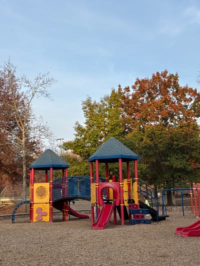A playground with a multi-tiered play structure stands on gravel. The structure features red posts, yellow panels, and blue roofs, with slides and climbing features. Trees in fall colors surround the playground, contrasting with the blue sky above. Some trees are bare, while others still have leaves in shades of orange, red, and green. The picture is taken from a low angle.