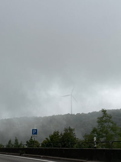 The image depicts an overcast and foggy landscape in the vicinity of Wiesensteig, Germany. The upper portion of the frame is dominated by a uniform grey sky and dense atmospheric fog. In the midground, a line of densely forested hills or mountains extends horizontally, largely obscured by the thick haze. A large, three-bladed wind turbine, characterized by a tall mast and three distinct blades, is partially visible above the tree line on the distant elevation, its form softened by the fog.

In the foreground, the edge of a paved road or highway occupies the lower section of the image, showing a dark asphalt surface with a single white lane marking. A dark grey concrete barrier or retaining wall separates the road from the adjacent green trees and bushes, which are more distinct than the distant forest but still partially enveloped in mist. On the left side, a blue rectangular road sign with a white 'P' symbol and the text "500m" is affixed to a pole. To the right, a white and black delineator post is positioned along the barrier. No individuals, animals, or vehicles are discernible in the scene. The overall environment suggests cool, damp, and low-visibility weather conditions.
