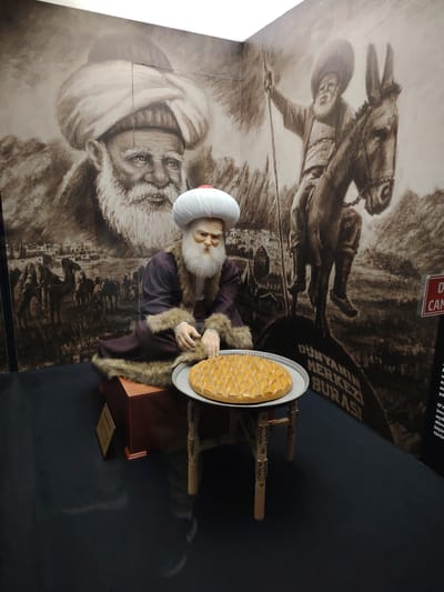 This image captures an indoor museum exhibit, likely in Eskisehir, Turkey, featuring a life-sized figure against a large wall mural. The central subject is a mannequin of an elderly man with a long, flowing white beard and mustache, wearing a prominent white turban with a red top and a traditional dark brown, fur-lined robe. He is seated on a wooden block, gazing thoughtfully at a round plate of diamond-patterned pastry, resembling baklava, which rests on a small, intricately carved wooden table in front of him.
The expansive, sepia-toned mural behind the figure depicts two main scenes: a large, detailed portrait of an old man with a white beard and turban on the left, closely resembling the mannequin; and on the right, a similar elderly man riding a donkey while holding a staff. The mural's background features mountains, a line of camels, and a distant settlement, evoking a historical and rural atmosphere.
Prominent text visible on the mural reads "DÜNYANIN MERKEZİ BURASI," which translates to "THE CENTER OF THE WORLD IS HERE," a quote famously attributed to Nasreddin Hodja. A partial red sign on the far right edge of the frame shows "Dİ..." and "CAN...". The dark, reflective floor completes the exhibit setting. The overall display pays homage to a significant historical or cultural figure, very likely Nasreddin Hodja, given the location context and common iconography.