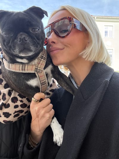 This close-up, low-angle image captures an affectionate moment between a woman and her dog in Strömstad, Sweden, during bright daylight.
The main subjects are a blonde woman and a black dog. The woman has short, light blonde hair and is wearing tortoise-shell framed sunglasses. She has a gentle smile on her face, with her head leaning against the dog's. She is dressed in a dark, possibly black, coat. Her right hand, seen holding the dog, sports a simple gold band ring and a delicate gold hand chain bracelet.
The dog is primarily black with distinctive white patches on its chest and paws, suggesting a breed like a French Bulldog or Pug mix. It wears a beige plaid harness, which has a pattern similar to Burberry. The dog appears calm and looks directly towards the viewer. Part of its body is covered by a soft, leopard-print fabric.
The background reveals an outdoor setting under a bright, possibly clear or partly cloudy sky. A light-colored building with windows is visible, suggesting an urban or town environment. The overall mood of the image is warm and loving, highlighting the bond between the woman and her pet. No visible text is present in the image.