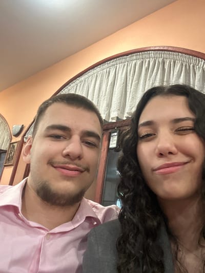 The image depicts two young adults, a male and a female, positioned in a close-up, chest-up shot within an indoor setting. The male, on the left, has short brown hair, light stubble on his chin and upper lip, and wears a light pink collared shirt. He exhibits a faint smile. The female, on the right, has long dark curly hair, wears a dark grey jacket, and has her right eye partially closed with a slight smirk. The background features light peach-colored walls, dark brown arched architectural elements, and light-colored gathered curtains. A wall sconce with a white cover is visible on the far left, above a dark framed picture or relief sculpture. Through an opening on the right, a dark object with faintly discernible white text, possibly reading "OPEN," is visible. The scene is illuminated by warm, artificial interior lighting. The specific location in the city of Arkovna, Bulgaria, cannot be determined from the visual information presented in the image.