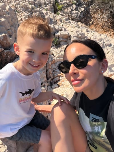 A woman and a young boy are posing for a photograph on a rocky surface. The woman is wearing sunglasses and a black t-shirt with a colorful graphic. The boy is wearing a white t-shirt with a shark graphic and patterned grey pants. They are both smiling. The background is a rocky landscape, possibly a beach. The location is Kostrena, Croatia.