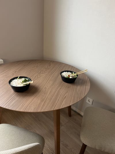 The image shows a high-angle view of a round, wooden table in a room with light-colored walls. Two black bowls of noodles with chopsticks are placed on the table. Two chairs with light-colored, textured upholstery are positioned near the table. The floor appears to be wooden. An electrical outlet is visible on the wall. The image's location is not possible to determine from the given information.