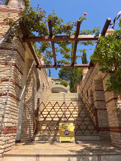 A stone stairway with a wooden trellis above it leads to a gated entrance to the Monastery of Archangel Michael, located at 40.597 latitude and 24.701 longitude in Moni Archaggelou, Greece. The trellis is covered in green vines with a few pink roses blooming.  A yellow sign reads "PLEASE DO NOT GO BEYOND THIS POINT." The stairway is made of stone steps with a light brown colored wooden gate.  The gate is closed. The surrounding area is filled with green trees and lush vegetation. The sky is blue and clear. The stone wall is made of rough, uneven stones.  The wall is also covered in green vines and foliage.