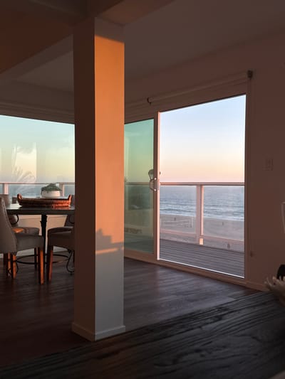 The image depicts an interior view of a residence situated in Manhattan Beach, United States, overlooking the Pacific Ocean at either dawn or dusk. The scene is illuminated by natural low-angle sunlight, casting warm orange-pink hues on interior surfaces and a large, white rectangular column prominent in the foreground. Dark hardwood flooring extends across the visible floor area. To the left, a dining area is visible, featuring a dark-topped table surrounded by at least three upholstered light-colored dining chairs. A woven basket holding a white planter with green foliage rests on the table.

Large windows and a partially open sliding glass door occupy the background, providing an expansive view of the exterior. Immediately beyond the sliding door is an outdoor deck with dark horizontal planking and a clear glass railing supported by white vertical posts. Beyond the deck, a wide, sandy beach is visible, featuring a white volleyball net with poles. The Pacific Ocean extends to the horizon, displaying gentle waves and reflecting the warm sky colors. The sky transitions from a pale blue overhead to a soft orange-pink near the horizon. No individuals or active subjects are present in the frame.