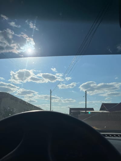 The image provides a view from inside a vehicle, looking through the windshield. The sky is a clear blue with scattered white clouds. Sunlight is visible, casting a bright glare. Several power lines stretch across the sky. In the distance, houses with red roofs and a corrugated metal fence are visible. A dark dashboard and steering wheel are in the foreground, partially obscuring the view. The image appears to have been taken in Telenești, Moldova, based on the context provided. There are no people or specific actions depicted in the scene. The windshield shows several cracks.