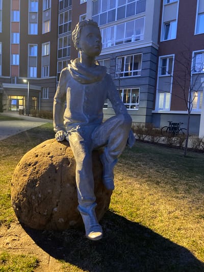 This media file captures an outdoor nighttime scene in Chemoshur, Russia, centered on a statue of a person resting on a large rock. The statue, crafted from a light grey, metallic material, depicts a young man or boy lying on his back, propped up on an elbow, with his head slightly elevated and looking upwards. He is dressed in casual attire, possibly a hooded top and trousers. The large, textured rock supporting him is brown.

The foreground and mid-ground feature grassy terrain, distinctly illuminated by an unseen artificial light source, which casts warm, yellowish light on some areas and plunges others into deep shadow. In the background, modern multi-story residential buildings are visible, their numerous windows reflecting or emitting cool, bluish-white light from inside. Bare tree branches are scattered throughout the scene. A bicycle rack, holding a bicycle, is discernible in the mid-ground near the buildings.

The time of day is evening or night, indicated by the prominent artificial lighting and the overall dark ambient conditions. The scene evokes a quiet, contemplative atmosphere, with the statue appearing to be in a state of peaceful rest. There is no specific activity taking place beyond the static depiction of the sculpture. No text is visible in the image.