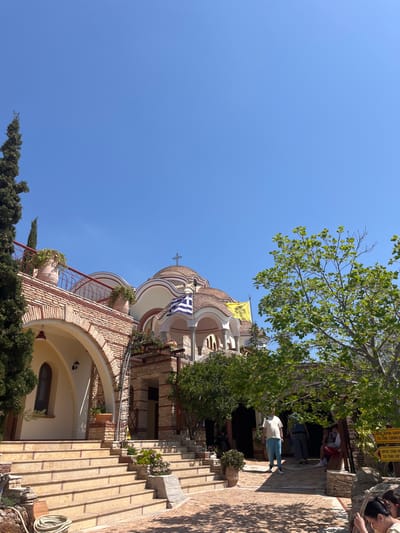 The image shows a view of the Monastery of the Archangel Michael in Moni Archaggelou, Greece (located at latitude: 40.597, longitude: 24.701). The Monastery is built in a traditional Greek architectural style with a mix of stone and stucco. The building is adorned with arches, columns, and a cross atop the dome. A Greek flag and a yellow flag are flying from the building. Several people are walking around the Monastery, while others are seated on steps or benches. There are trees, greenery, and flowers surrounding the Monastery. The sky is bright blue and clear.  The ground is made of stone pavers. A sign in Greek is visible in the lower right corner. The image is taken from a low angle, showing the Monastery from the ground level.