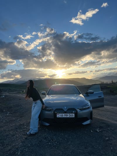A woman leans against the front of a gray BMW with an open driver-side door. The vehicle's license plate reads "66 MH 066" with "AM" indicating Armenia. The background shows a setting sun behind cloudy sky over a hilly landscape. The location is possibly near Hrazdan, Armenia.