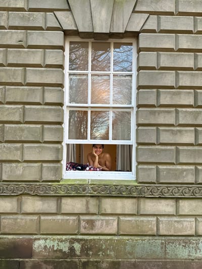 An exterior photograph depicts a person looking out from a window of a stone building in Stafford, United Kingdom. The building facade features light-colored, rusticated ashlar block masonry. A white, multi-paned sash window, with its lower sash open, is centrally framed. Above the window, a classical stone architrave with a keystone-like element is visible, and below the windowsill, a carved stone molding displays a continuous scroll pattern. The lower section of the wall exhibits green discoloration consistent with biological growth.
Inside the open window, a smiling individual with dark hair is visible from the upper torso, appearing bare-chested. Their right cheek rests on their right hand, with the elbow positioned on the windowsill. A red and black plaid fabric is draped along the interior windowsill. Light-colored curtains are present on either side of the interior window frame. The upper panes of the window reflect bare tree branches against an overcast sky, indicating the external environment.