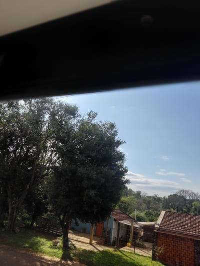 This image depicts a view of a residential area in Encarnacion, Paraguay, from an elevated perspective, possibly from a window. The foreground is dominated by a thick, dark, horizontal object, likely the interior frame of a window or sunroof, casting a shadow that partially obscures the upper portion of the scene.
Below this obstruction, the natural environment comes into focus. A large, leafy tree with a dense canopy is prominently featured on the left side of the image, its branches extending upwards and partially into the sunlight. The tree's dark foliage contrasts with the bright, clear blue sky, which is dotted with a few wispy white clouds.
Further into the scene, beyond the large tree, a cluster of buildings is visible. These structures appear to be houses or small outbuildings. One building has a light blue wall and a reddish-brown tiled roof. Another building to its right has a more prominent reddish-brown tiled roof, constructed with brick walls. A fence, possibly made of metal or wood, separates parts of the property.
The ground in the foreground and mid-ground is covered with green grass and dirt paths, with sunlight casting dappled shadows from the trees. The overall impression is of a sunny day in a semi-rural or suburban setting. The presence of residential buildings and dense vegetation suggests a typical neighborhood.