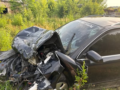 The image shows a severely damaged black car, likely the result of a car accident. The front of the vehicle is heavily crumpled, with the hood and bumper mangled. The windshield is cracked extensively, and a portion of the car's engine is exposed. A white sticker with text is visible on the upper part of the windshield. The car is situated in an outdoor setting with tall green grass and trees surrounding it. In the background, there's a glimpse of a brick building and some industrial-looking structures, suggesting a rural or semi-industrial area. The time of day appears to be daytime, with natural light illuminating the scene. The overall impression is one of destruction and abandonment, possibly after an accident. There are no people visible in the image. The text on the windshield appears to be "memento mori."