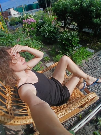 In an outdoor setting, a woman with short, curly brown hair is resting on a wicker chair. She is wearing a black tank top and shorts. Her right hand is raised to her forehead, and her eyes are closed. The chair is positioned on a gravel pathway. Behind her, a lush garden is visible, featuring various plants and flowers, as well as a greenhouse and a fence. The scene suggests a relaxed moment in a garden, likely in Maslova Pristan', Russia, given the typical rural landscape.