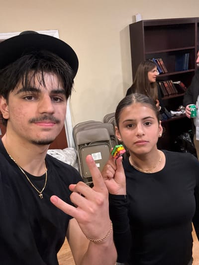 The image depicts an indoor scene in Bergenfield, United States, featuring four individuals. In the foreground, a young adult male with dark hair, a mustache, wearing a black hat, black t-shirt, gold chain necklace with a pendant, and a gold chain bracelet on his right wrist, poses with his right hand forming a "rock on" gesture. To his right, a young adult female with dark hair pulled back, wearing a black long-sleeved shirt, layered gold necklaces, and small stud earrings, holds a small, rectangular, multi-layered confection (colored green, yellow, pink, and red) in her right hand. Both foreground subjects are looking directly at the camera.
In the mid-ground, a dark brown wooden bookshelf filled with books, some appearing to be religious texts, stands against a light-colored wall. Stacked beige plastic and metal folding chairs are visible between the foreground subjects and the bookshelf. Two additional adult individuals are partially visible in the mid-ground: a female with dark hair appears to be in conversation near the bookshelf, and on the far right, a male's right hand holds a green soda can. The environment suggests a communal or informal gathering space.