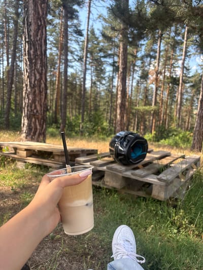 The image depicts a hand holding a clear plastic cup filled with a layered beverage, likely iced coffee. A black straw extends from the cup. The setting is a forest, characterized by tall, slender pine trees and dry grass. In the background, several wooden pallets are stacked, and a black motorcycle helmet with blue accents rests on one of them. The scene appears to be outdoors, in a natural environment. While the provided location is Chavdar, Bulgaria, the image itself does not contain any explicit visual cues to confirm or deny this specific geographical detail.