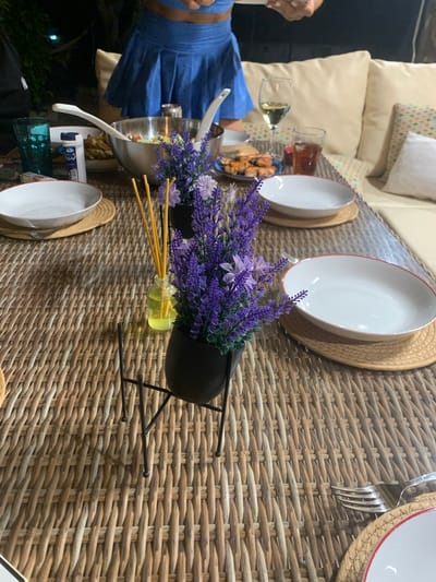 This media file depicts an outdoor dining scene, possibly in Lloseta, Spain, at dusk. A woven table holds plates, cutlery, and glasses. In the foreground, a small bouquet of lavender in a black planter sits beside a bottle with yellow liquid and reeds, suggesting a diffuser. A person, partially visible and wearing a blue dress, is seated at the table. The setting appears to be a patio or garden, with hints of cushions and greenery in the background. The lighting suggests it's evening.