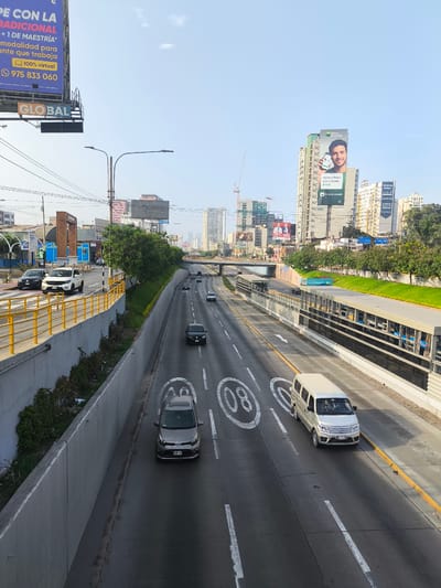 The image depicts a highway in La Victoria, Peru. The road has multiple lanes in each direction and is filled with various vehicles including cars, a van, and a truck. Overhead road signs are visible, including the painted "80" speed limit. On both sides of the highway, there are concrete barriers topped with greenery. There is an elevated bus rapid transit (BRT) system on one side of the highway. Tall buildings and billboards rise up in the background. A large billboard with a man is visible, as well as another one with text in Spanish advertising a 100% virtual master's degree program. The sky is light blue and mostly clear.