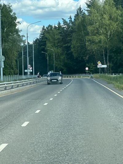 The image shows a gray Mini Cooper driving on a paved road with dashed white lane markings. The car is in the center of the frame, facing forward. The road appears to be a highway or a major road, bordered by a metal guardrail on the left and trees on both sides. The sky is cloudy, with patches of blue. Several streetlights are visible along the left side of the road. There are traffic signs visible on the right side of the road, including a diamond-shaped sign with two cars and an arrow, and a circular sign with a red border and a horizontal bar. The location is identified as Lukovnya, Russia. The overall scene depicts a sunny day on a road surrounded by nature.