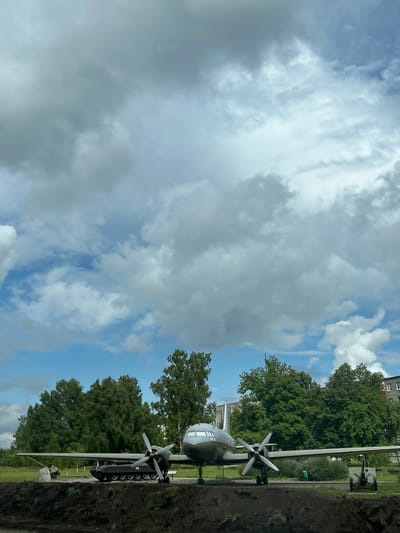 An external, eye-level, full shot captures an old, propeller-driven airplane displayed outdoors, likely as a monument, against a backdrop of lush green trees and a cloudy sky. The aircraft, a light gray color with white-tipped red propellers, is positioned on a slightly elevated mound of dark soil in the foreground. A dark colored tank is visible behind the airplane's left wing, and a small artillery piece is situated behind the right wing. To the right of the airplane, a multi-story building with a red roof is partially visible, suggesting an urban or semi-urban setting within the city of Бягомльскі сельскі Савет, Belarus. There are no people visible in the frame. The overall ambiance is one of historical preservation and outdoor exhibition.