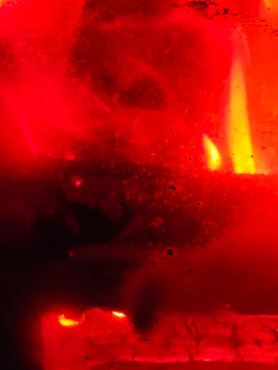 A close-up shot reveals a vibrant red and orange scene, possibly a fire within an enclosure. A faint yellow flame flickers at the upper right. Dark, indistinct forms exist within the blurred red space. Small, irregular spots dot the surface, adding texture. The bottom section displays linear elements that suggest structures, glowing red.