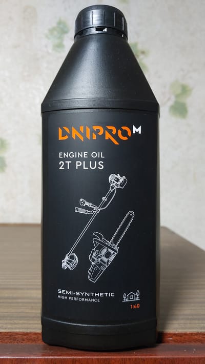A black plastic bottle of "DNIPROM ENGINE OIL 2T PLUS" is positioned on a wooden surface. The bottle has an orange logo and white text indicating the product type and specifications. A line drawing of a string trimmer and a chainsaw is printed on the bottle. Below this, "SEMI-SYNTHETIC HIGH PERFORMANCE" is written in white text, along with icons representing a house and trees, and a ratio of "1:40". The background is a textured, light-colored wall with a subtle pattern. This scene is located in Oleśnica, Poland.