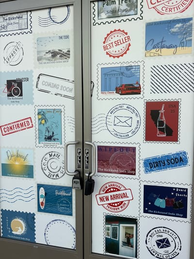 The image captures a glass door, likely of a business in Huntington Beach, USA, adorned with a collage of graphics designed to resemble postage stamps. The door is bisected horizontally by a metal frame that includes silver pull handles, each labeled "PULL" and "DOOR," with a black Master combination lock secured to the right handle.
The numerous "stamps" on the door advertise "The Kickstand Soda Shop" and feature various coastal-themed designs and products. Visible text on these stamps includes names such as "Palm Tree Soda Shop," "THE TIDE," "BEST SELLER," "Castaway," "CRUISER," "Lemon Breeze," "Daybreak," "Dirty Classic," "WIPEOUT," "Paddleboard," and "surf sup." Promotional phrases like "COMING SOON," "NEW ARRIVAL," "APPROVED," "CONFIRMED," and "CERTIFIED" are also present. Several stamps include postal-related text like "NORTH POLE POST," "MAIL," "POSTAL SERVICE CENTRAL OFFICE," and one shows "USA 25." The location "Newport Beach CA" is explicitly mentioned on multiple stamps, indicating a nearby coastal setting.
The stamp illustrations depict items such as a bicycle, a paddleboard, soda bottles, board shorts, a classic red vehicle, a lemon tree, a shipwreck, ocean waves, a sunrise, a palm tree, and an envelope. One stamp contains a small, framed photograph of individuals at a counter inside what appears to be the soda shop. The overall aesthetic suggests a casual, beach-oriented establishment, consistent with a coastal California location like Huntington Beach.