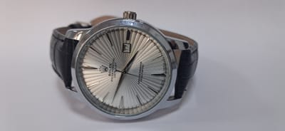 The image shows a close-up of a Rolex Oyster Perpetual Datejust watch with a silver, radially patterned dial and a black leather strap. The watch is positioned on a white, neutral surface, with the casing and strap prominently visible. The time appears to be approximately 1:51, with the date showing "17". The watch is the sole subject of the image, and there are no people or other objects present. The background is a plain white surface, suggesting a studio or controlled environment for product photography. The lighting is soft and diffused, highlighting the details of the watch. The image is taken from a slightly elevated, side angle, focusing on the watch face and strap. There is no indication of the city of Danghara, Tajikistan, in the image or its context.