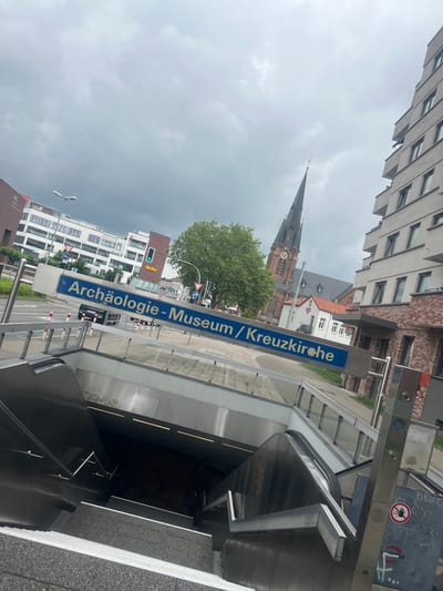 This media file captures an urban street scene in Herne, Germany, under an overcast sky. The image is oriented vertically but appears to be rotated 90 degrees clockwise, showing elements as if viewed from an unusual angle.
In the foreground, a modern subway (U-Bahn) entrance, characterized by metallic and dark surfaces, descends into the ground via an escalator. Near the entrance, various metal structures display graffiti, and a red circular sign with a crossed-out white hand symbol (likely indicating "Do not touch" or "No access") is visible.
Dominating the mid-ground is a large blue sign with white German text, clearly reading "Archäologie-Museum / Kreuzkirche." This sign points towards the locations of an archaeological museum and a church.
Behind the sign, to the left, a street is visible with a green traffic light, suggesting active vehicle traffic. Several multi-story buildings with white facades and numerous windows line the street. A red sign for the "Netto" supermarket is identifiable on one of these buildings.
To the right, behind the sign and partially obscured by a green tree, stands a historic church with a prominent, dark spire and a visible clock face, built with traditional brickwork. This church is likely the "Kreuzkirche" mentioned on the blue sign.
The upper portion of the image reveals more modern buildings with grey and brick facades and many windows, blending into the cloudy, uniformly grey sky. The overall impression is a bustling urban area with a mix of historical and contemporary architecture on an overcast day.