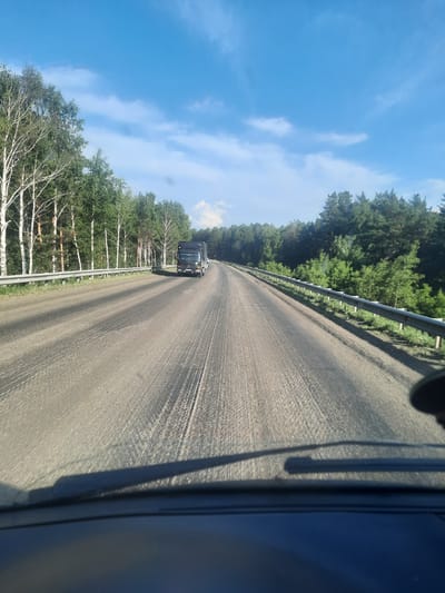 The image shows a perspective from inside a vehicle looking out at a road lined with trees. A black truck is visible driving on the road in the distance. The sky is blue with scattered white clouds, suggesting a sunny day. The road appears to be paved and has visible tire tracks. On either side of the road, there are dense green trees, with some birch trees with white trunks visible closer to the foreground on the left. A metal guardrail runs along the side of the road. The overall scene depicts a journey through a wooded area on a clear day. The perspective suggests the photograph was taken while the vehicle was in motion. No text is visible in the image.
