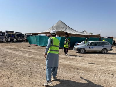 The image depicts an unpaved outdoor worksite in Pir Dust, Afghanistan, under a clear blue sky. The ground is dry, light brown dirt and gravel with visible vehicle tracks. In the foreground, a male subject, viewed from the back, wears a light blue shalwar kameez, a lime green high-visibility vest, and a white hard hat, walking towards the midground.
In the midground, several large, light-colored canvas tents are erected, featuring green tarpaulin sections as partial walls or enclosures. To the left of these tents, a line of multiple dark-colored heavy-duty dump trucks is parked. To the right of the tents, a silver Toyota Corolla station wagon with a blue "GCD" logo on its side is parked. A white sedan is partially visible behind the station wagon.
Three additional individuals are observed in the midground. One male, wearing a dark top, a lime green high-visibility vest, and a white hard hat, walks towards the right side of the frame. Another male, in similar attire, stands near the white sedan. A person is visible within the driver's seat of the silver station wagon. The scene is illuminated by daylight, casting distinct shadows.