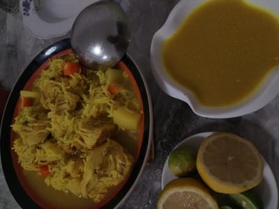 A vibrant yellow dish of rice and meat with potatoes and carrots sits in a dark-rimmed bowl, accompanied by a large silver spoon. Nearby, a white dish contains a smooth, yellow sauce. A smaller white bowl holds lemon and lime halves and wedges. The setting appears to be a casual dining area. The image suggests a flavorful, possibly Middle Eastern or Asian-inspired meal.