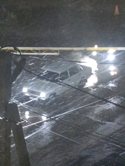 A silver car is illuminated by its headlights in a heavy downpour. Water covers the road, reflecting lights above. Power lines crisscross the foreground, obscuring the view of the car. To the left, a utility pole is visible. A yellow barrier stretches horizontally across the scene, and an orange traffic cone stands to the upper right. The overall scene is dark and conveys hazardous driving conditions.