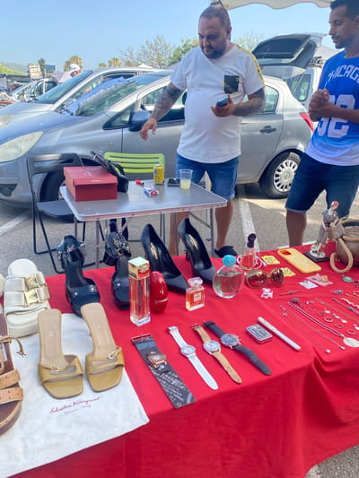 This image depicts an outdoor market scene, likely a car boot sale or flea market, with a bright red tablecloth covering a table laden with various items for sale. The setting appears to be San Luis De Sabinillas, Spain, indicated by the presence of palm trees and typical Mediterranean architecture in the background. Two men are present; one, wearing a white t-shirt with yellow trim and blue denim shorts, is holding a smartphone. The other, in a blue t-shirt with white lettering, stands beside him. The table displays a variety of merchandise, including women's footwear such as high heels and sandals, perfume bottles, sunglasses, jewelry including necklaces and rings, watches, a lighter, and a yellow smartphone case. Behind the table, a silver car with its trunk open is partially visible, suggesting a car boot sale setup. The ground is asphalt, and the lighting indicates a sunny day.