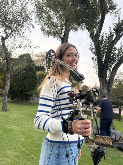 A woman holds a camouflage bow and arrow outdoors. She wears a white sweater with blue stripes and blue jeans. A man is visible in the background, seemingly involved with archery equipment. Trees and grass surround the scene, indicating a park or outdoor shooting range. The sky is partly cloudy.