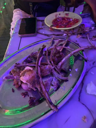 The image displays a close-up, high-angle view of a table after a meal, bathed in distinctive purple and blue artificial lighting. The central subject is a large, clear glass or plastic platter laden with numerous gnawed meat bones, primarily ribs, suggesting consumption of grilled or roasted meat. The platter's rim reflects a vivid green glow from the ambient light.
To the left, a small, light-colored bowl holds a salad of diced red onions and tomatoes, characteristic of "kachumbari," a popular Kenyan accompaniment. Scattered on the light-colored table, which has a marble-like pattern with dark veins, are pieces of white bread or flatbread. A dark, rectangular object, possibly a smartphone or power bank, rests on the table beside the salad bowl.
A person's arm and shoulder are partially visible on the far left, indicating someone is present at the table. The dominant purple and blue lighting, complemented by green reflections, creates a distinct nighttime or indoor atmospheric setting, likely a restaurant, bar, or social venue. The type of food remnants, particularly the grilled bones and kachumbari, aligns with culinary practices found in Kimorori, Kenya. No visible text is present. The scene portrays the remnants of a recently enjoyed communal meal.